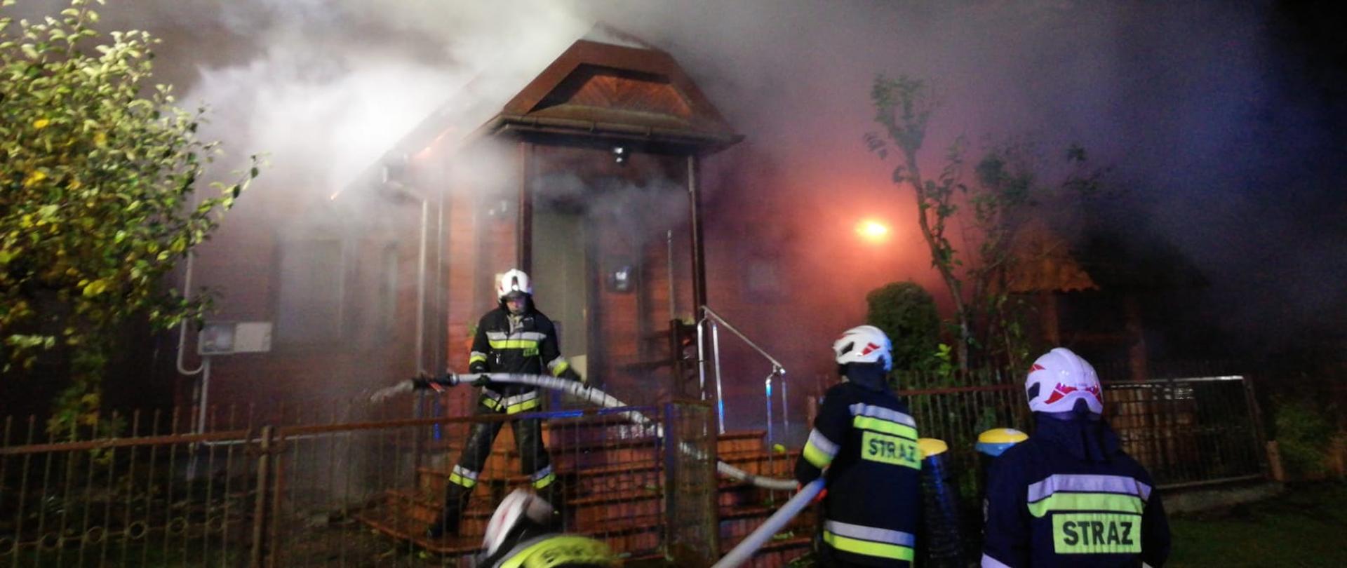na zdjęciu widać strażaków z prądem wody z zewnątrz budynku, z budynku wychodzi dym