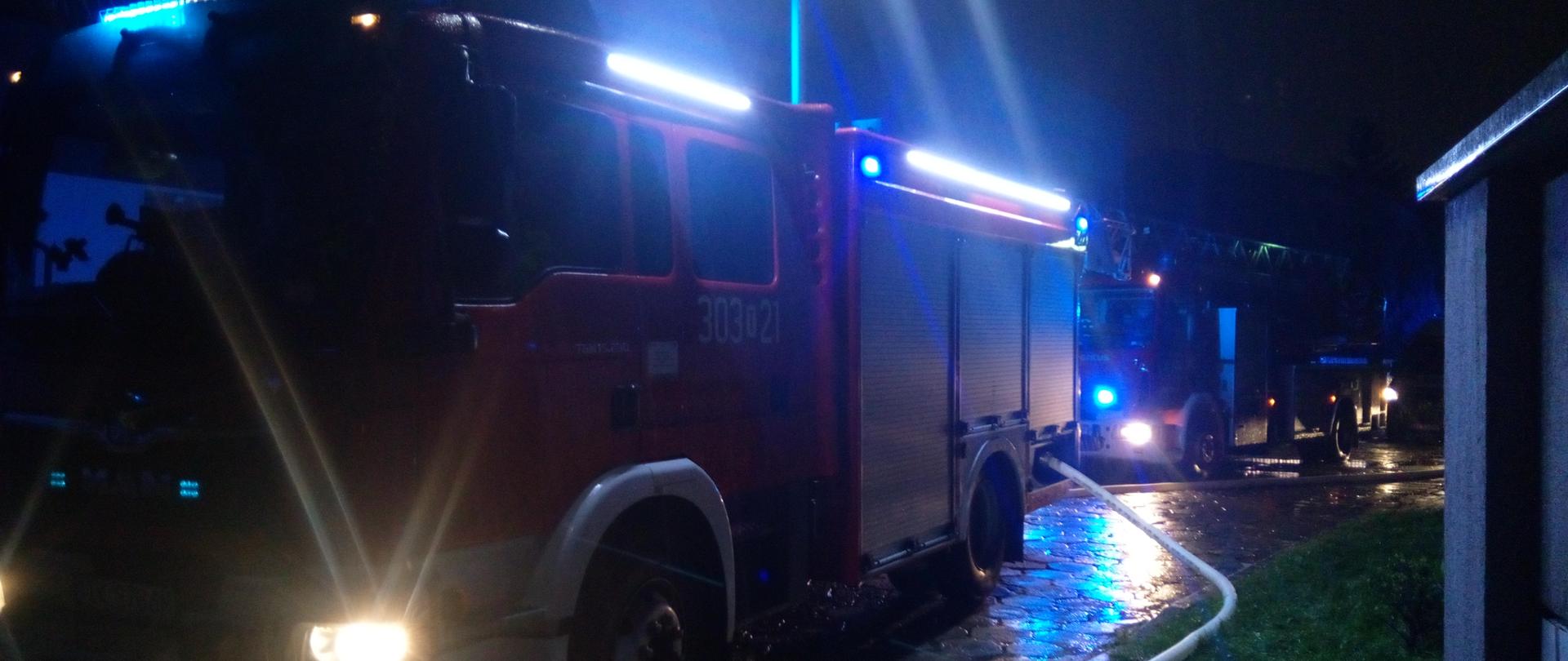 Zdjęcie przedstawia samochody strażackie oraz rozwinięte linie gaśnicze. Na zewnątrz jest ciemno i deszczowo. Wokół samochodów widać niebieską poświatę od sygnałów świetlnych. 