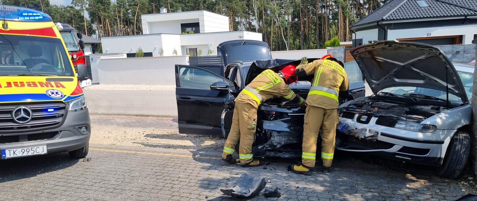 Zdjęcie przedstawia dwa uszkodzone pojazdy blokujące częściowo drogę powiatową. Na zdjęciu widać strażaków, którzy pracują przy odłączaniu akumulatorów w obu pojazdach. Po lewej stronie stoi zaparkowany pojazdy karetki pogotowia z włączonymi błyskowymi światłami alarmowymi w kolorze niebieskim.