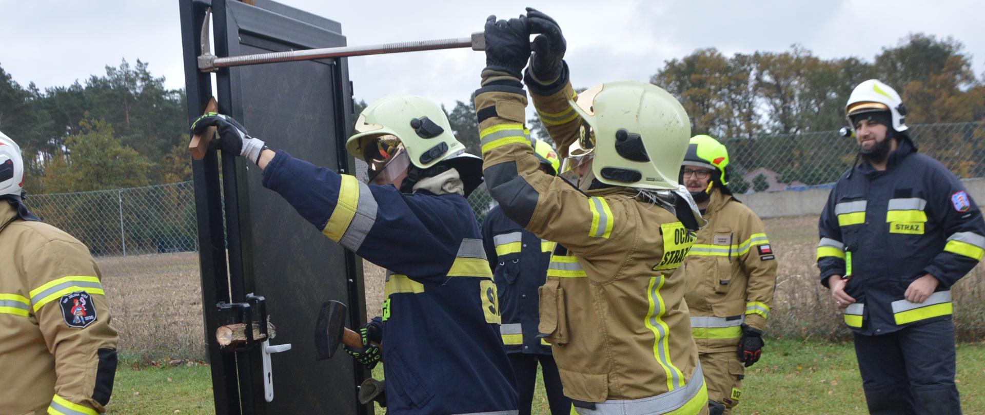 Warszaty szkoleniowe. Zdjęcie przedstawia fragment stadionu w Sarnowie, na którym przygotowane jest stanowisko do "siłowego otwierania drzwi". Na pierwszym planie ustawione sa specjalne drzwi do ćwiczeń, przy których znajduje się dwóch strażaków ratowników OSP. Przy pomocy sprzętu burzącego podejmuja próbę ich otwarcia. Jeden z nich ma ubranie specjalne koloru granatowego, drugi - piaskowego. Ich dziąlaniom przypatrują się inni strażacy. W tle rosną drzewa. 