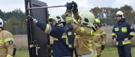 Warszaty szkoleniowe. Zdjęcie przedstawia fragment stadionu w Sarnowie, na którym przygotowane jest stanowisko do "siłowego otwierania drzwi". Na pierwszym planie ustawione sa specjalne drzwi do ćwiczeń, przy których znajduje się dwóch strażaków ratowników OSP. Przy pomocy sprzętu burzącego podejmuja próbę ich otwarcia. Jeden z nich ma ubranie specjalne koloru granatowego, drugi - piaskowego. Ich dziąlaniom przypatrują się inni strażacy. W tle rosną drzewa. 