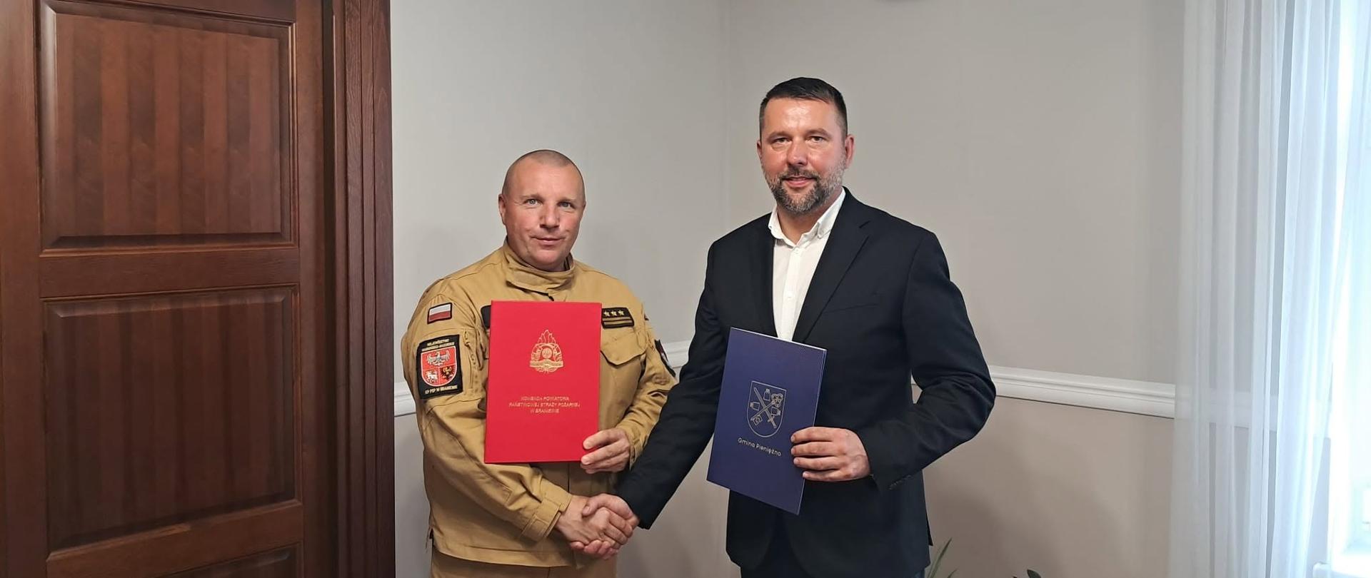 Na zdjęciu burmistrz Pieniężna oraz komendant powiatowy straż pożarnej, ściskają sobie dłonie oraz trzymają teczki.