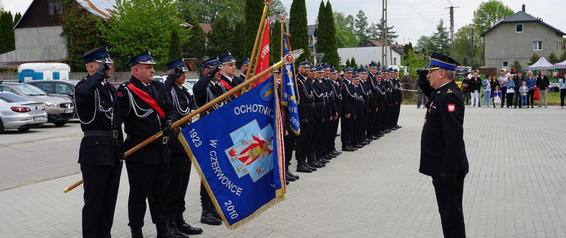 Zastępca Mazowieckiego Komendanta Wojewódzkiego PSP brygadier Waldemar Wysowski wita się ze sztandarem jednostki OSP w Czerwonce poprzez salutowanie. Poczet sztandarowy salutuje sztandarem poprzez jego pochylenie. Na dalszym planie strażacy stojący w szyku i zgromadzone osoby cywilne. 