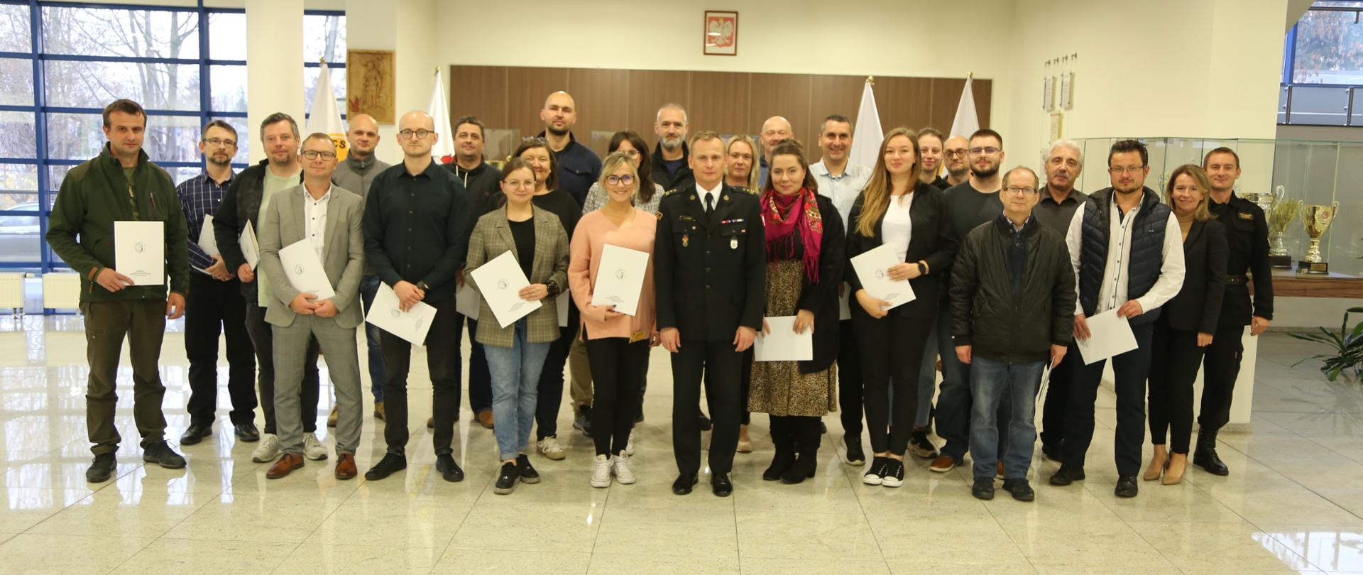 Grupowe zdjęcie uczestników szkolenia (24 osoby) w towarzystwie Zastępcy Komendanta Centralnej Szkoły PSP, funkcjonariusza CS PSP oraz kobiety. Uczestnicy szkolenia trzymają otrzymane zaświadczenia