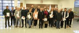 Grupowe zdjęcie uczestników szkolenia (24 osoby) w towarzystwie Zastępcy Komendanta Centralnej Szkoły PSP, funkcjonariusza CS PSP oraz kobiety. Uczestnicy szkolenia trzymają otrzymane zaświadczenia