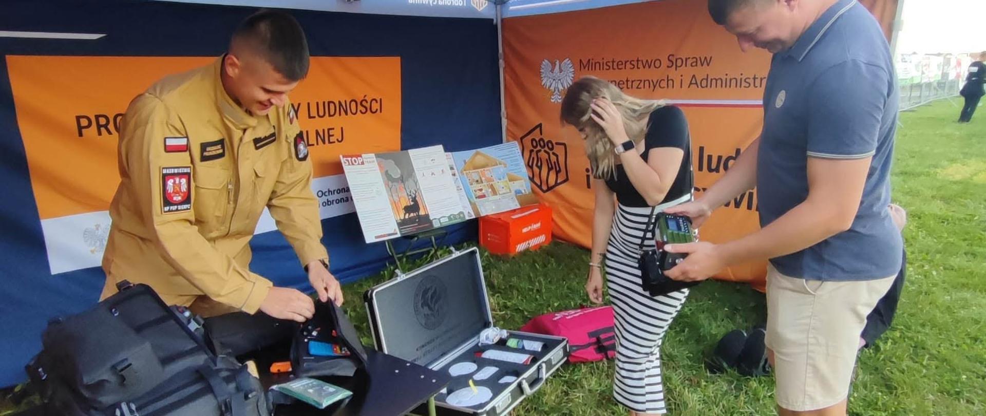Strażak w piaskowym mundurze pakuje sprzęt na stole pod niebiesko-pomarańczowym namiotem MSWiA, podczas gdy obok kobieta w paski i mężczyzna oglądają jedno z urządzeń.