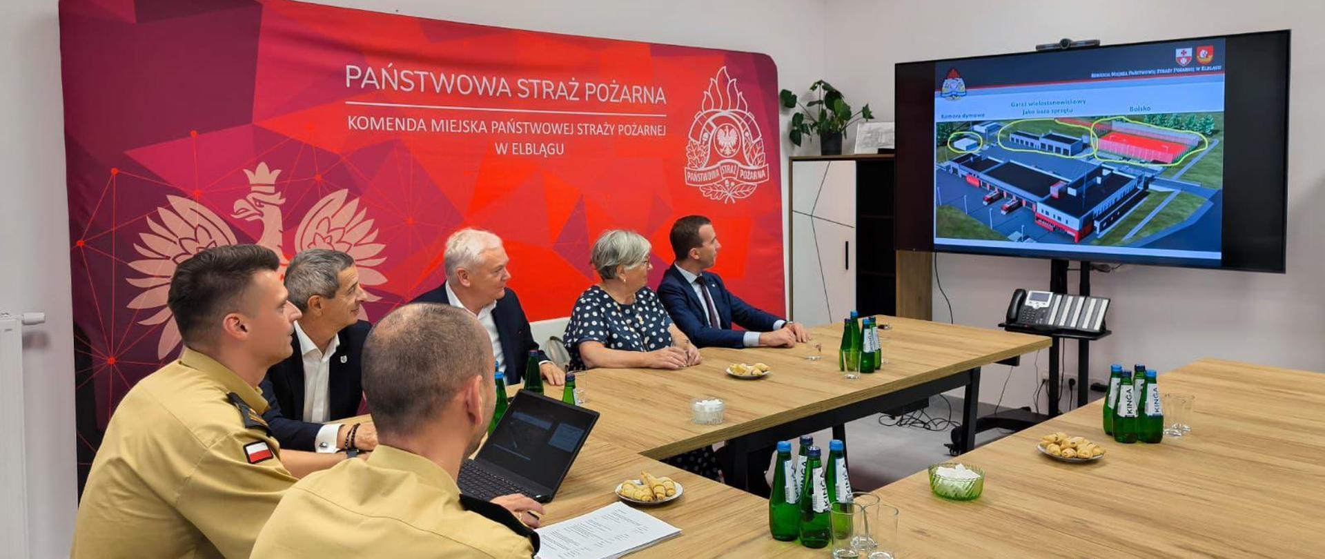 Na zdjęciu znajduje się grupa osób w sali odpraw Komendy Miejskiej Państwowej Straży Pożarnej w Elblągu. Osoby siedzą przy stole i oglądają i zapoznają się z prezentacją planu rozbudowy elbląskiej komendy. Prezentację przedstawia Komendant Miejski PSP w Elblągu. Wśród zgromadzonych osób jest Warmińsko - Mazurski Komendant Wojewódzki PSP oraz parlamentarzyści wchodzący w skład Żuławskiego Zespołu Parlamentarnego.