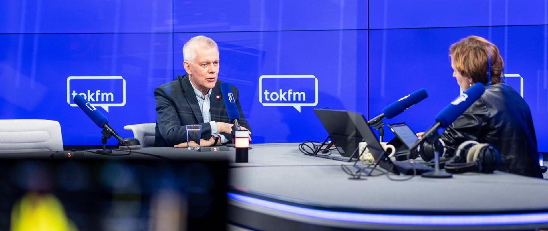 Na zdjęciu wnętrze studia radiowego. Przy szarym stole, na białych krzesłach, przed niebieskimi mikrofonami TOK FM siedzą - minister Tomasz Siemoniak (po lewej) oraz redaktor Karolina Lewicka (po prawej). Na stole sprzęt radiowy m.in. słuchawki. W tle niebieska ścianka wizyjna z białym logo TOK FM.