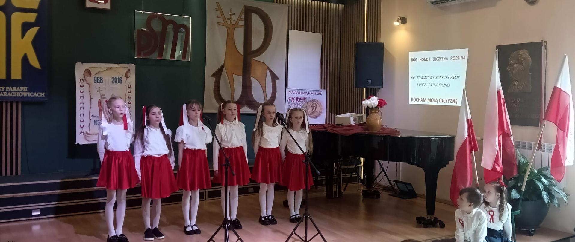 Na zdjęciu widać grupę młodych dziewczynek stojących na scenie i śpiewających. Ubrane są w białe bluzki i czerwone spódnice, a niektóre mają czerwone kokardy we włosach. Przed nimi ustawione są mikrofony na statywach. W tle znajduje się fortepian oraz ekran z wyświetlonym napisem: „BÓG HONOR OJCZYZNA RODZINA XXIII POWIATOWY KONKURS PIEŚNI I POEZJI PATRIOTYCZNEJ KOCHAM MOJĄ OJCZYZNĘ”. Scena ozdobiona jest patriotycznymi symbolami, takimi jak polskie flagi i historyczne plakaty. Po prawej stronie, przy scenie, siedzi dwójka dzieci w białych strojach z biało-czerwonymi kotylionami. Widzowie na sali oglądają występ, a jedna z osób nagrywa go telefonem. Pomieszczenie jest jasne, z eleganckim drewnianym wykończeniem i zielonymi ścianami.