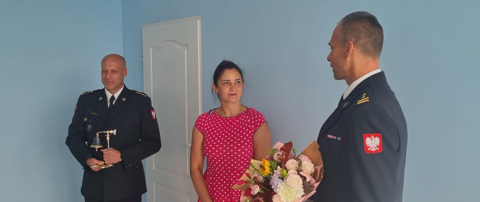 Zdjęcie przedstawia 3 osoby, z czego 2 to strażacy, a trzecia to kobieta w różowej sukience w białe kropki. Komendant Powiatowy PSP przekazuje bukiet kwiatów dla żony odchodzącego na emeryturę strażaka