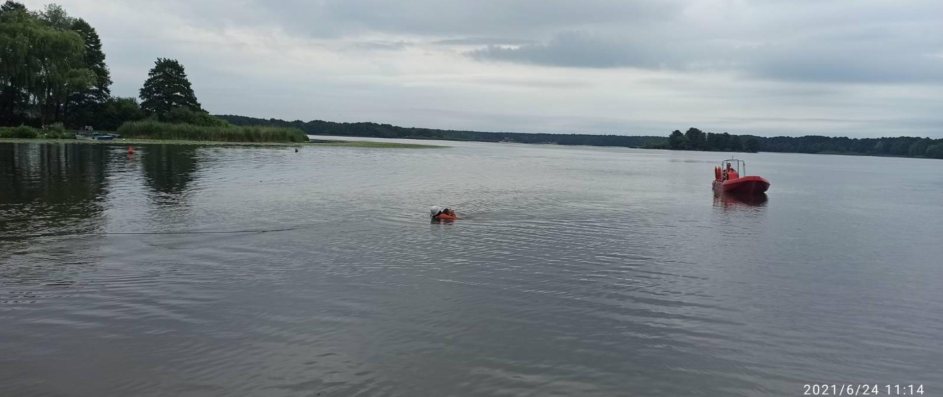 Zdjęcie przedstawia taflę jeziora, po której pływa czerwona łódź. Po lewej stronie od niej w wodzie znajduje się człowiek w pomarańczowym kombinezonie, który zabezpiecza osobę podtopioną