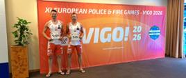 Daj strażacy stoją w pomieszczeniu przed wielką czerwoną ścianką imprezy z jasnym napisem „VIGO! 2026”. Mają na sobie sportowe koszulki z godłem Polski i spodenki oraz klapki. W rękach trzymają imienne numerki startowe do akredytacji: numer 2420 (Marcin Melnarowicz) oraz numer 2416 (Bartłomiej Baniak).