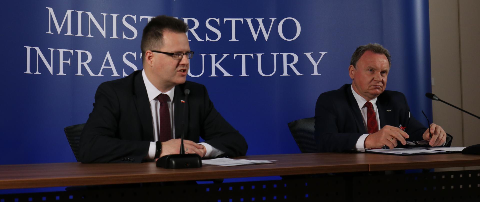 Na zdjęciu widać dwóch mężczyzn siedzących przy stole, w ich tle widać niebieską ściankę z białym napisem Ministerstwo Infrastruktury