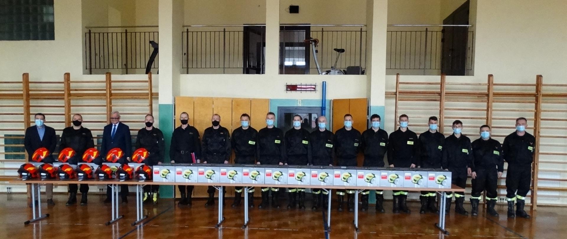 Zdjęcie zrobione na sali gimnastycznej w szeregu stoją strażacy ze zmiany służbowej w ubraniach dowódczo sztabowych, kierownictwo komendy w ubraniach dowódczo sztabowych, starosta jasielski Adam Pawluś, Prezes Lotos Infrastruktura w Jaśle, Jacek Gogola, wszyscy w maseczkach, przed strażakami stoi stół a na nim leżą hełmy strażackie, część hełmów znajduje się w pudełkach