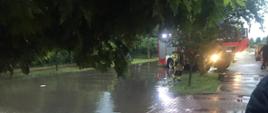 Na zdjęciu w porze wieczornej widać zalaną ulicę na skutek ulewnego deszczu w Ostrowcu Św. W tle samochód strażacki stojący w wodzie oraz strażak.