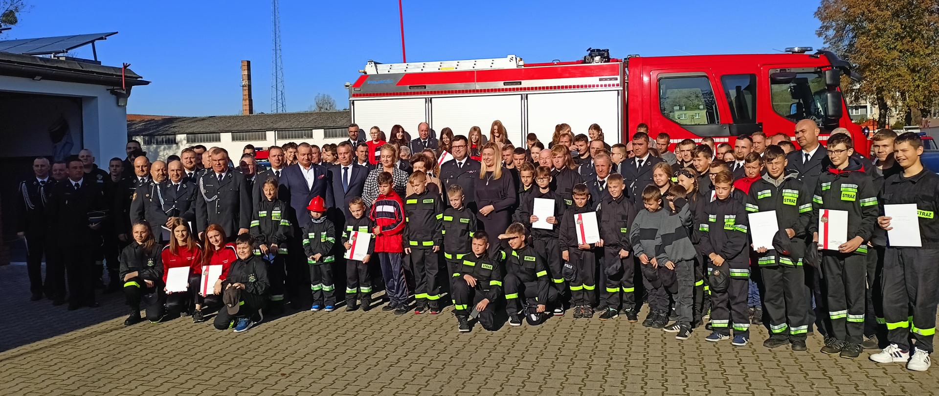 Zdjęcie przedstawia wspólne zdjęcie grupowe wszystkich uczestników uroczystej zbiórki na tle samochodu pożarniczego OSP Kuklinów