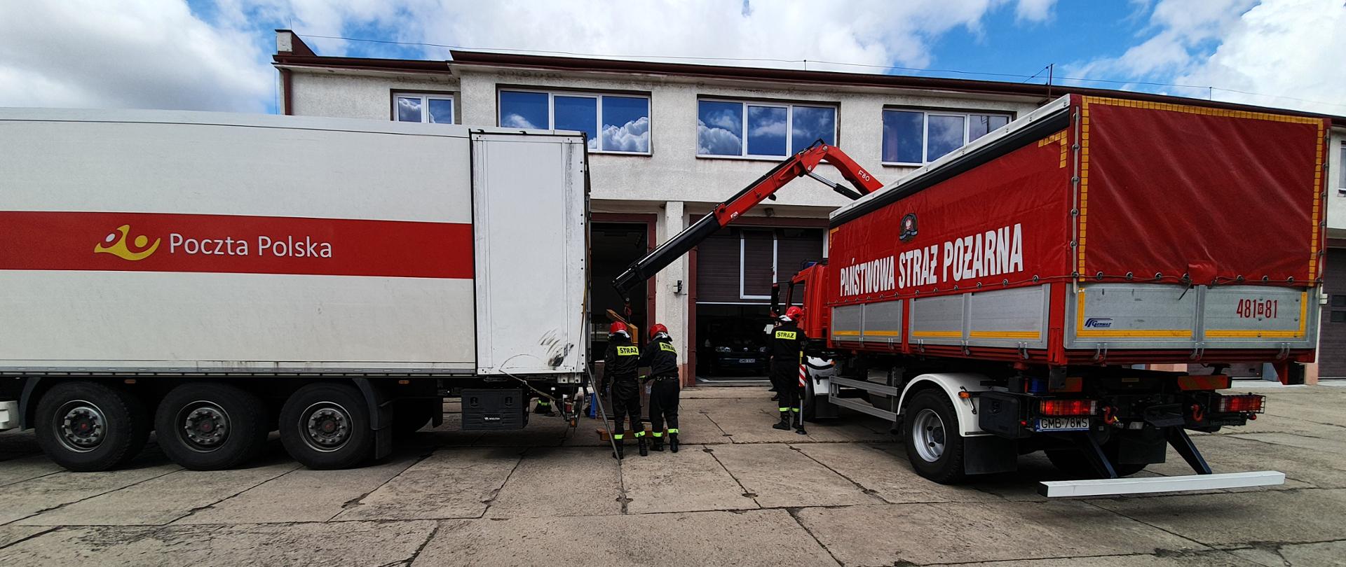 Fotografia przedstawia etap rozładunku samochodu poczty polskiej przez strażaków. Dwa samochody ciężarowe stoją obok siebie przed budynkiem Komendy Powiatowej Państwowej Straży Pożarnej w Malborku.