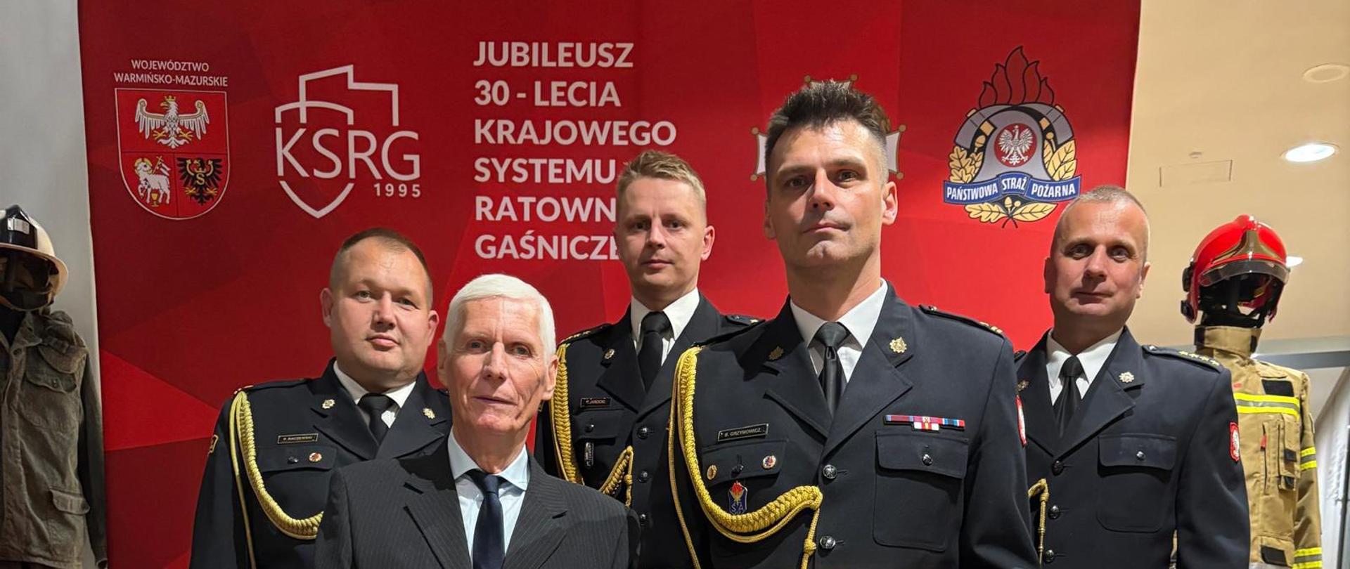 Funkcjonariusze Państwowej Straży Pożarnej ubrani w mundury galowe, pozują do zdjęcia. W ręku trzymają dyplom. W tle za placami baner z napisem 30 lat krajowego systemu ratowniczo gaśniczego.