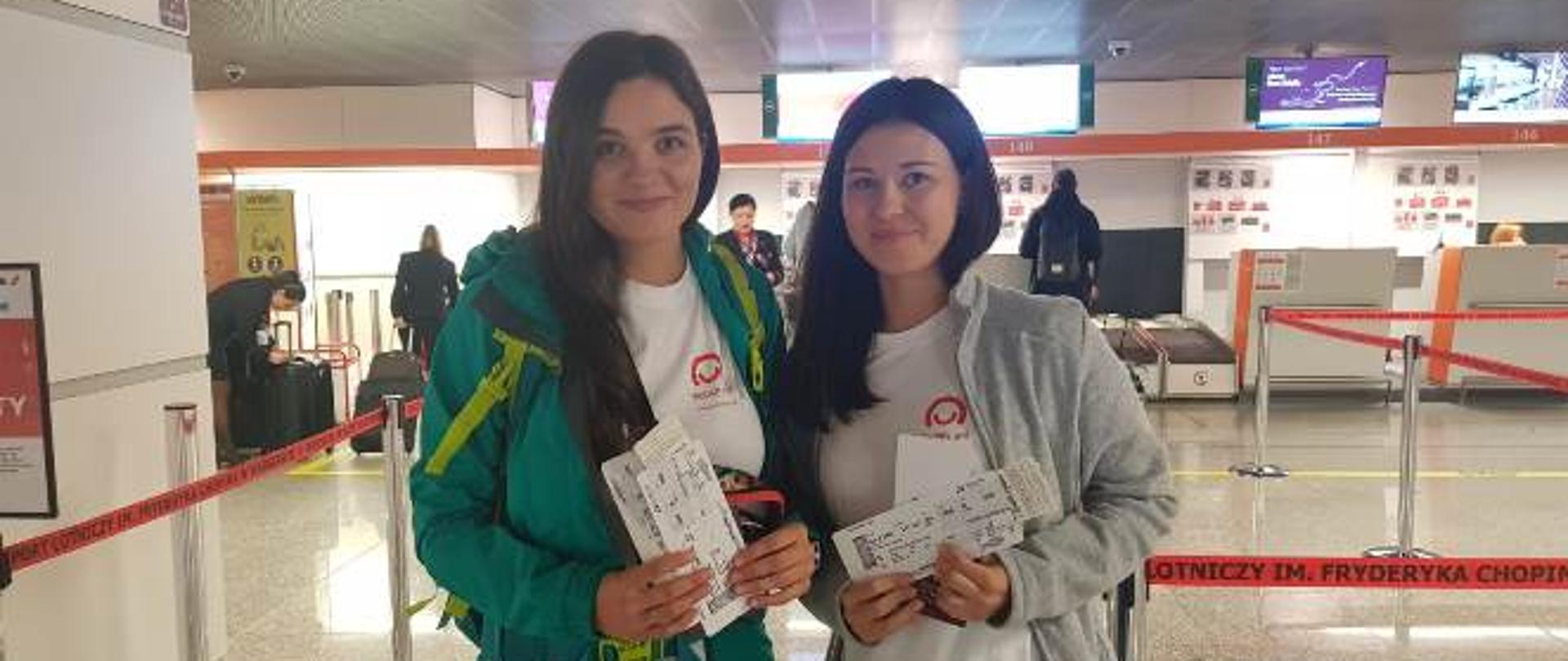 Dwie wolontariuszki w koszulkach Polskiej pomocy z biletami po odprawie na lotnisku Chopina w Warszawie