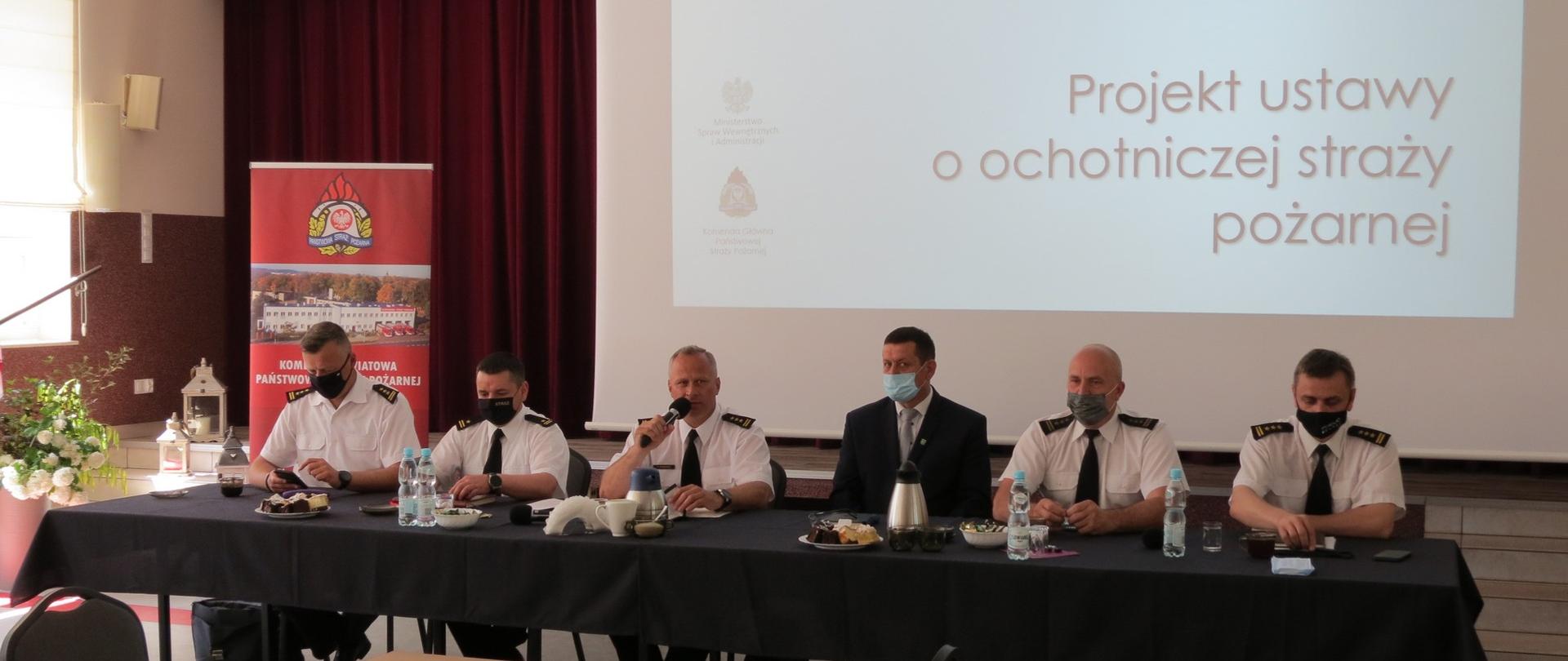 Widok na stół prezydialny, przy którym siedzą komendanci, rozmawiając z uczestnikami debaty, siedzącymi na na sali. Za ich placami ekran z wyświetlonym tekstem omawianej ustawy.