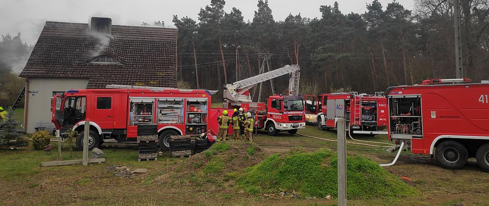 Na zdjęciu znajduje się budynek mieszkalny z którego wydobywa się dym oraz samochody straży pożarnej.