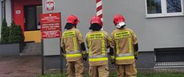 Trzech strażaków zawieszających flagę na maszcie przed budynkiem Straży Pożarnej.