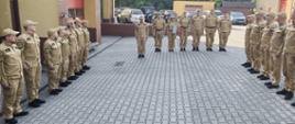 Grupa strażaków w piaskowych mundurach bojowych i czapkach z daszkiem stoi na wybrukowanym placu przed budynkiem remizy. Uformowali oni szyk w kształcie podkowy: dwa szeregi stoją naprzeciw siebie po bokach, a mniejsza grupa dowódców zamyka formację w głębi placu. Na zdjęciu widoczna jest oficjalna zbiórka strażaków na placu manewrowym przed budynkiem Państwowej Straży Pożarnej. Wszyscy zgromadzeni, czyli ponad dwudziestu strażaków, mają na sobie jednakowe, nowoczesne mundury w kolorze piaskowym oraz czapki z daszkiem. Stoją w zorganizowanym szyku przypominającym literę „U”. Po lewej i prawej stronie placu stoją dwa szeregi strażaków zwróconych do siebie twarzami. Plac wyłożony jest szarą kostką brukową. Po lewej stronie znajduje się fragment budynku remizy z ceglanymi elementami i czerwonymi bramami garażowymi. W tle, za plecami stojących w głębi strażaków, widać zaparkowane samochody osobowe oraz fragmenty innych budynków, a w oddali zielone drzewa. Pogoda jest słoneczna.