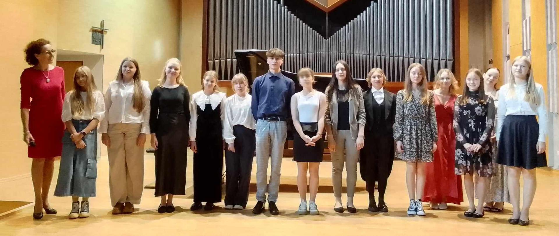 Sala koncertowa, wszyscy uczestnicy koncertu stoją na scenie, po ich lewej stronie pani prowadząca koncert, za nimi w tle organy.