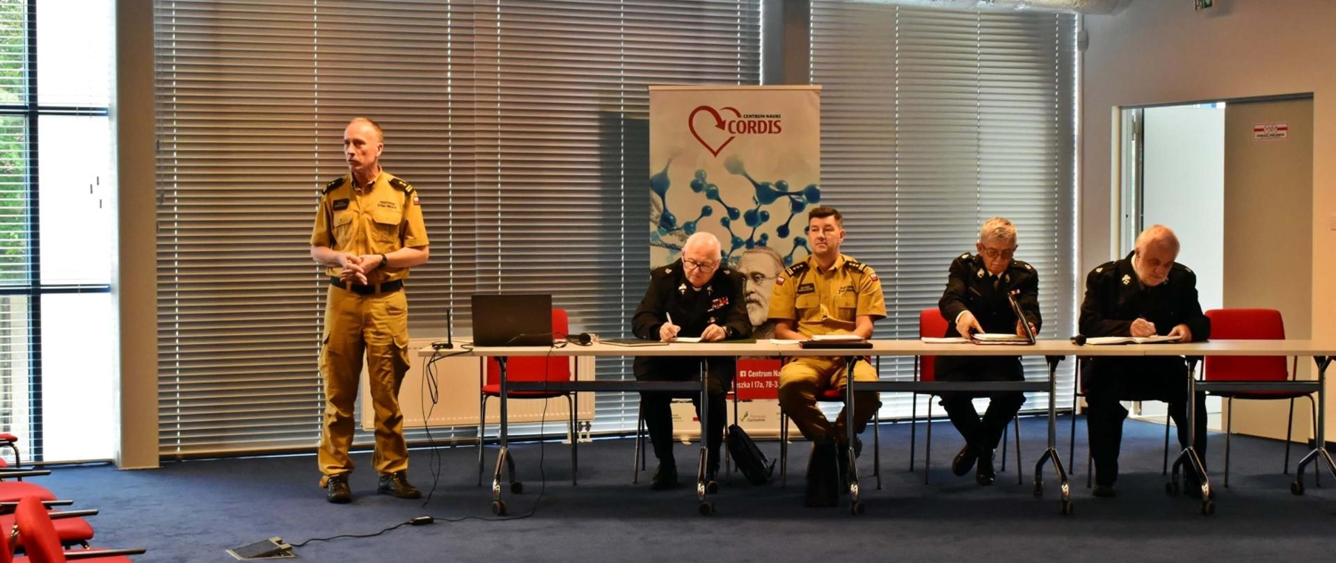 Zdjęcie przedstawia uczestników spotkania z druhów OSP z Zastępcą Zachodniopomorskiego Komendanta Wojewódzkiego PSP w centrum nauki CORDIS. Jedna osoba stoi i wygłasza przemówienie.