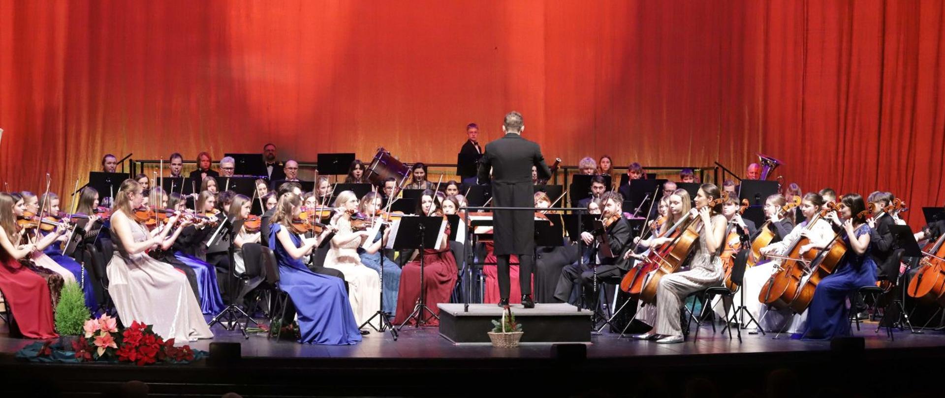 Na zdjęciu widać orkiestrę symfoniczną występującą na scenie Teatru Ziemi Rybnickiej. Muzycy siedzą w półkolu, grając na różnych instrumentach — widać skrzypce, wiolonczele, altówki oraz instrumenty dęte w dalszej części sceny. Na środku, tyłem do publiczności, stoi dyrygent w czarnym fraku, prowadzący orkiestrę.
Tło sceny stanowi ciepła, czerwono-pomarańczowa kurtyna, a oświetlenie nadaje całości elegancki, uroczysty nastrój. Muzycy są odświętnie ubrani — kobiety w długich, balowych sukniach w różnych kolorach, mężczyźni w garniturach lub frakach. Na przodzie sceny widać ozdobne kwiaty: cyprys w czarnej doniczce i gwiazdy betlejemskie w trzech kolorach.