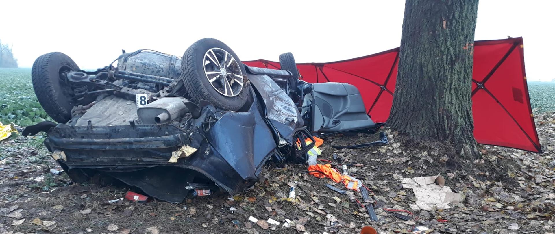 TRAGICZNY PORANEK. Na zdjęciu widać czerwony parawan, obok drzewa leży rozbity samochód osobowy, porozrzucane elementy, rzeczy.