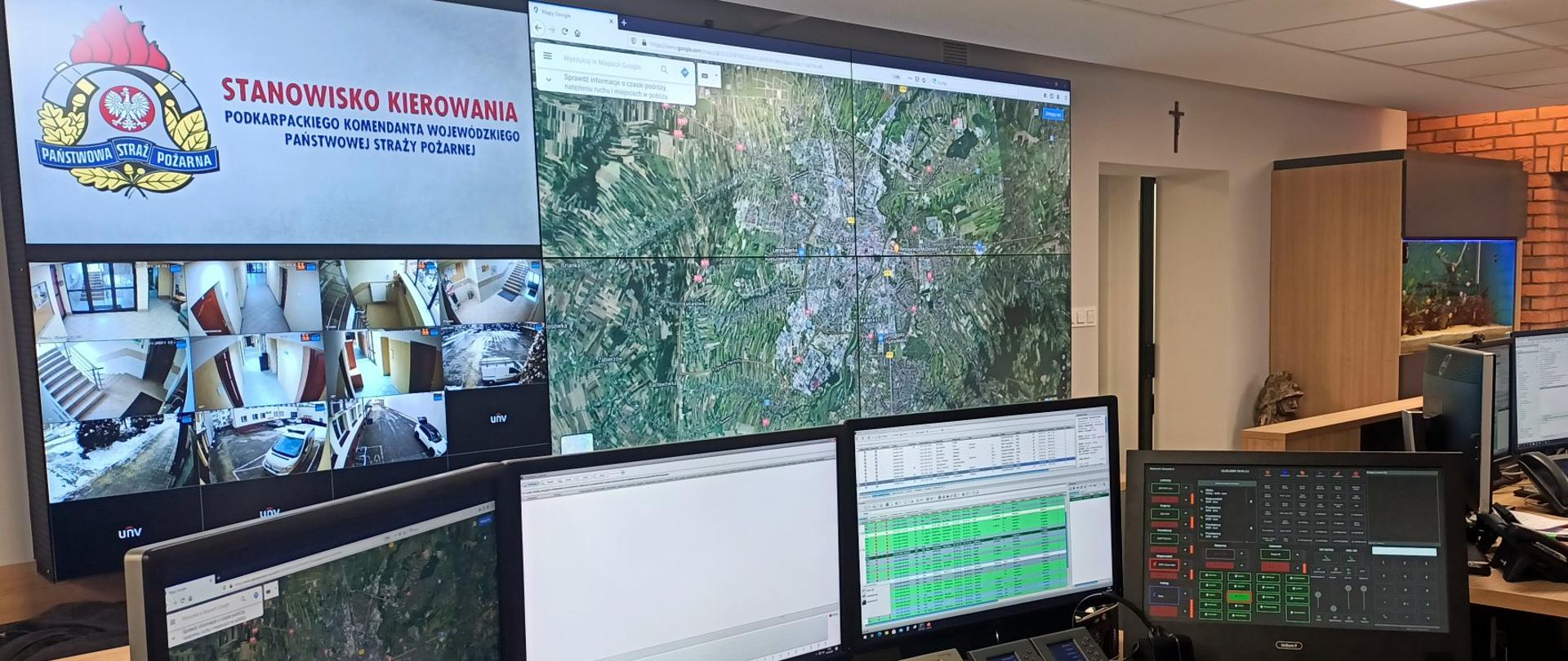 Stanowisko Kierowania Podkarpackiego Komendanta Wojewódzkiego PSP. Na pierwszym planie zdjęcia pokazane stanowisko pracy dyżurnego operacyjnego. Na biurku umieszczone są trzy monitory komputerowe, dwie centralki telefoniczne oraz, po lewej stronie stoi konsola dyspozytorska. W tle widać ścianę wizyjną, składającą się z sześciu monitorów, na których wyświetlona jest mapa satelitarna okolic Rzeszowa, widok z kamer monitoringu oraz Stanowisko kierowania Podkarpackiego Komendanta Wojewódzkiego PSP