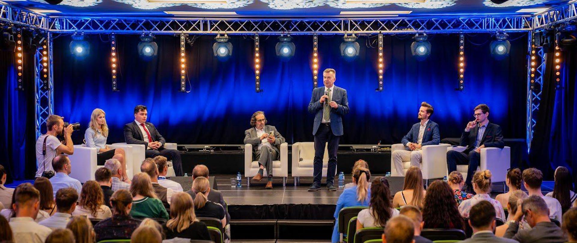 Przed salą wypełnioną siedzącymi na ustawionych w rzędy krzesłach ludźmi na oświetlonym na niebiesko podwyższeniu stoi minister Wieczorek i mówi do trzymanego w ręku mikrofonu, za nim na fotelach siedzi kilka osób.