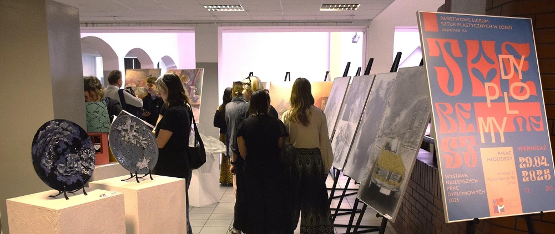 Na zdjęciu uczniowie podczas zwiedzania wystawy. Na pierwszym planie plakat promujący wydarzenie oraz ceramiczne talerze na kubikach