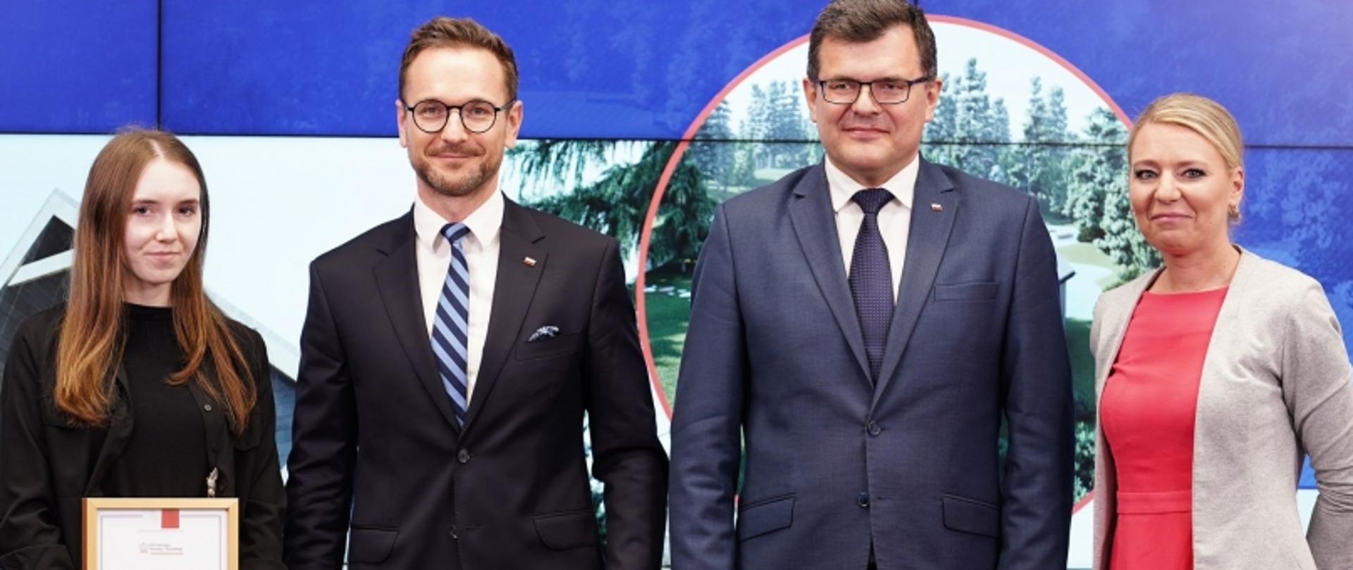 Grupa czterech osób w eleganckich strojach, jedna z nich trzyma oprawiony certyfikat; w tle ekran z obrazem plenerowym.