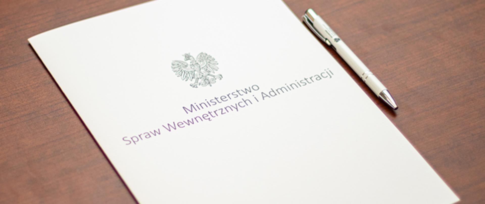 Na zdjęciu widać leżącą teczkę na dokumenty z napisem "Ministerstwo Spraw Wewnętrznych i Administracji" i orłem z godła narodowego i leżący długopis MSWiA.
