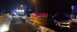 Zdjęcie wykonane w porze nocnej. Na zdjęciu widać jeden z pojazdów biorących udział w wypadku. Pojazd osobowy w kolorze szarym posiada rozbity przód. Dalej w pewnej odległości widać pojazd pożarniczy w kolorze czerwonym ze srebrnymi elementami zabudowy. Pojazd posiada włączoną świetlną sygnalizację alarmową w kolorze niebieskim. Na prawo od pojazdu znajduje się ustawiony czerwony parawan zasłaniający rozbity wrak jednego z pojazdów. Z lewej strony pojazdu pożarniczego stoi strażak ubrany w żółte ubrani specjalne i czerwony hełm. 