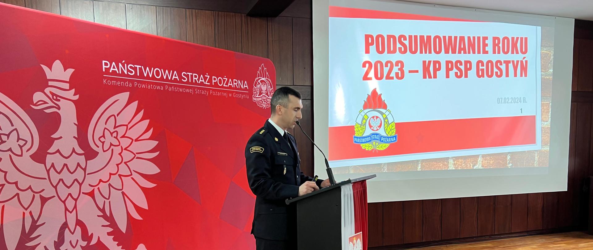 Komendant powiatowego w Gostyniu przedstawiającego prezentację. Stoi on przy mównicy i przemawia przez mikrofon. Za nim wyświetlana jest prezentacja na rzutniku.