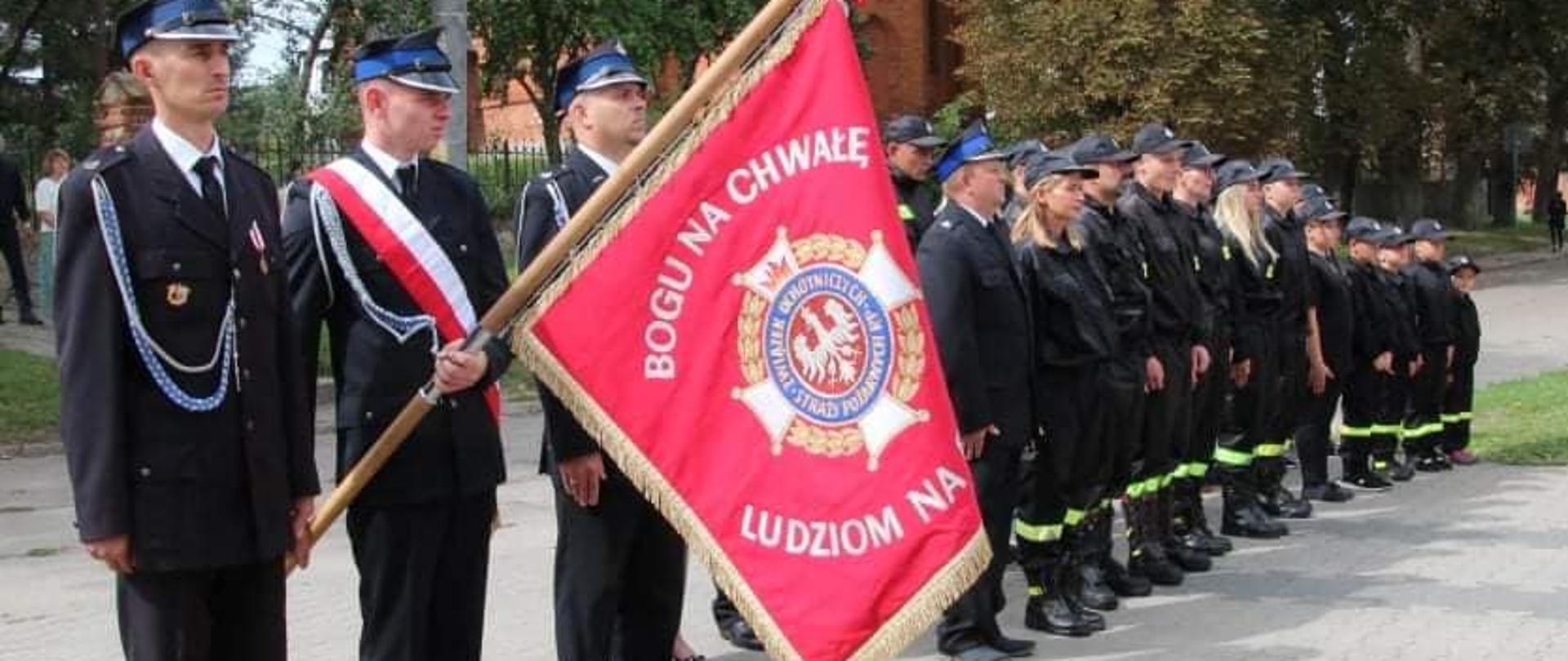 Na zdjęciu strażacy OSP w umundurowaniu stojący w szyku. Jeden z druhów prezentuje sztandar.