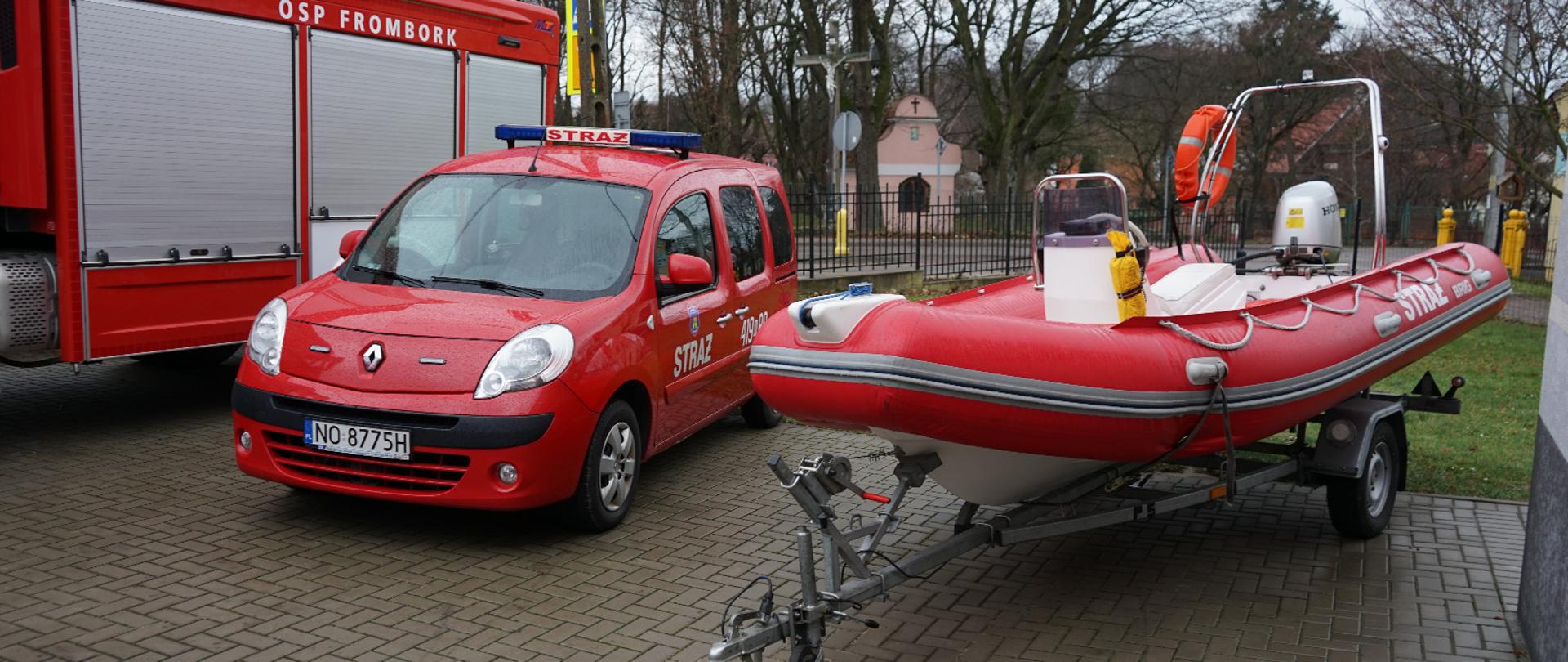 Na placu z kostki brukowej stoją z lewej strony dwa samochody pożarnicze, czerwone. Z prawej strony stoi czerwony ponton strażaki na przyczepie. W oddali drzewa.