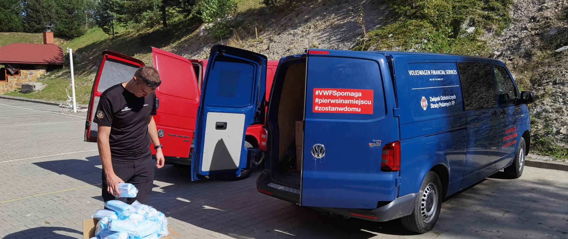 Zdjęcie przedstawia dwa samochody dostawcze na tle krajobrazu. Na pierwszym planie znajduje się strażak ładujący środki dezynfekcyjne do samochodu.