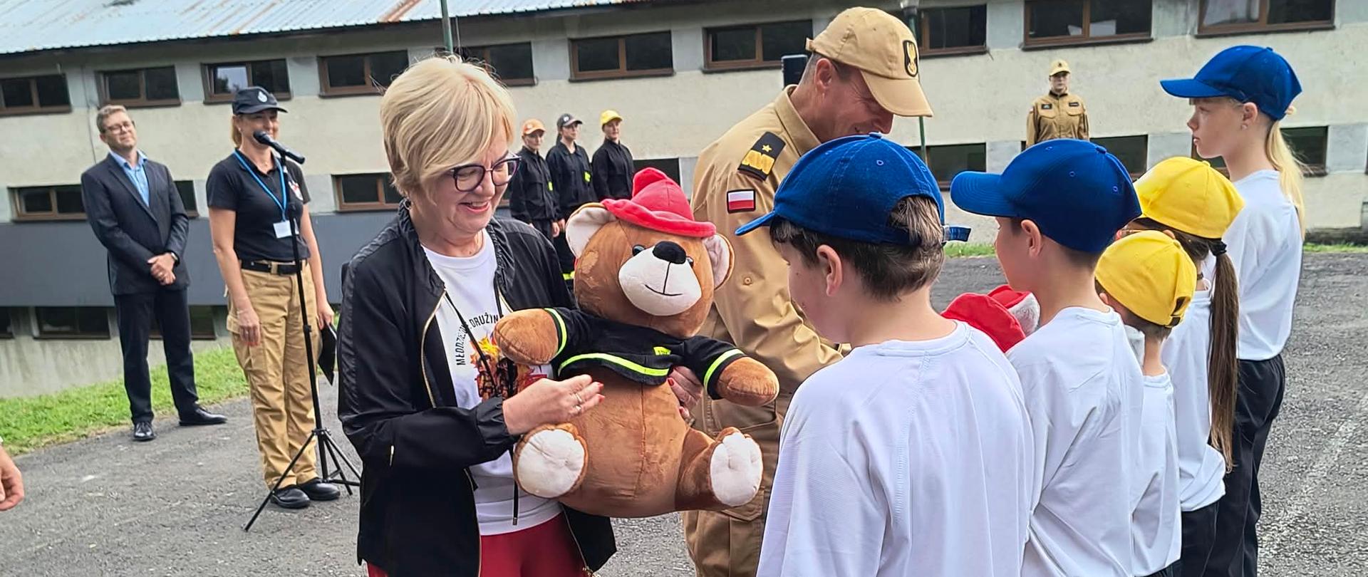 Pani Teresa Kubas-Hul, Wojewoda Podkarpacki, uśmiecha się, wręczając dużego pluszowego misia w strażackim mundurze i czerwonym kapeluszu chłopcu z Młodzieżowej Drużyny Pożarniczej podczas oficjalnej uroczystości na obozie szkoleniowym w Bieszczadach. Pani Wojewoda ma na sobie białą koszulkę, czarną kurtkę, czerwoną spódnicę i okulary. Chłopiec stoi w szeregu rówieśników w białych koszulkach, ciemnych spodniach i czapkach z daszkiem (niebieskich i żółtych), tyłem do obiektywu. Za nimi stoi strażak w piaskowym mundurze służbowym. W tle, na placu przed piętrowym budynkiem z dachem z blachy falistej, widać innych uczestników w mundurach strażackich oraz dorosłych opiekunów, w tym kobietę z mikrofonem przy statywie. Niebo jest zachmurzone. Zdjęcie wykonano z poziomu wzroku.