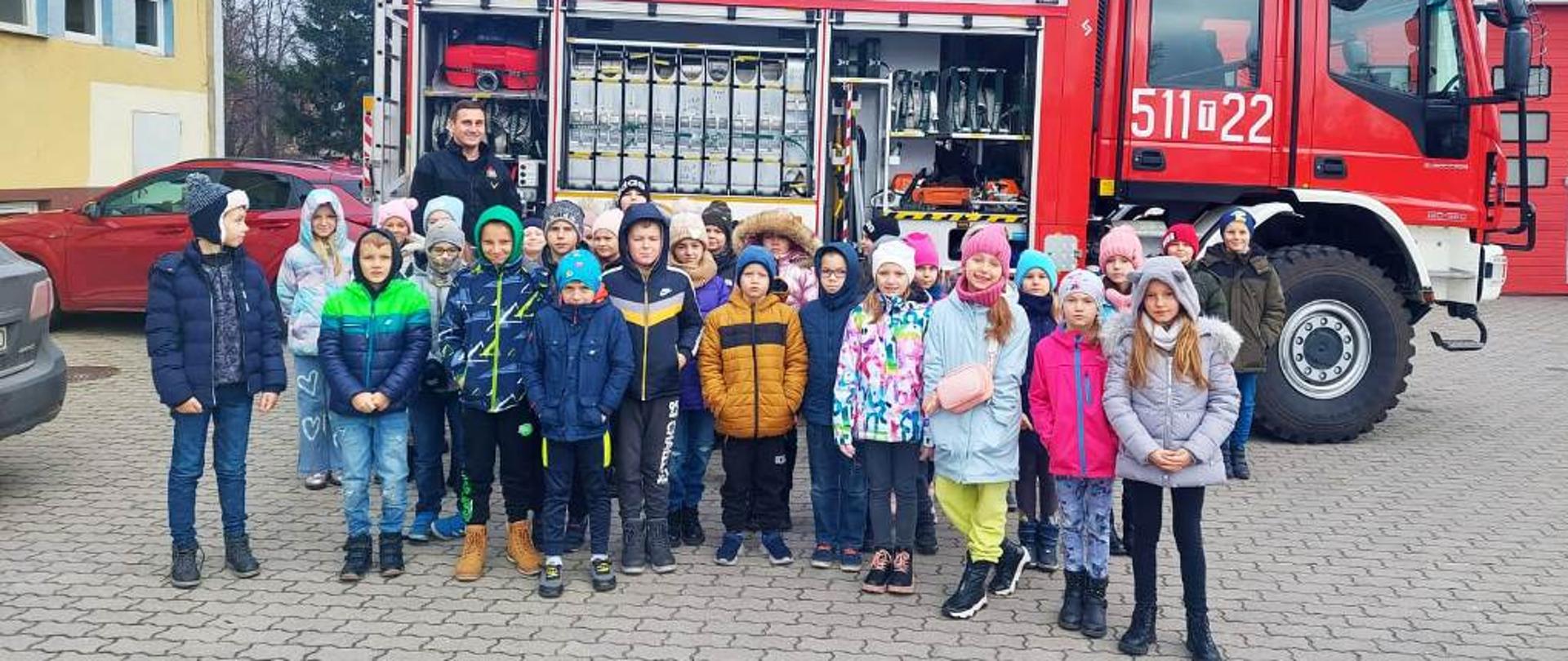 Plac wewnętrzny Komendy Powiatowej PSP we Włoszczowie, uczestnicy wycieczki, uczniowie szkoły podstawowej ustawieni przed samochodem ratowniczo-gaśniczym, samochód ma otwarte skrytki w których znajduje się sprzęt pożarniczy
