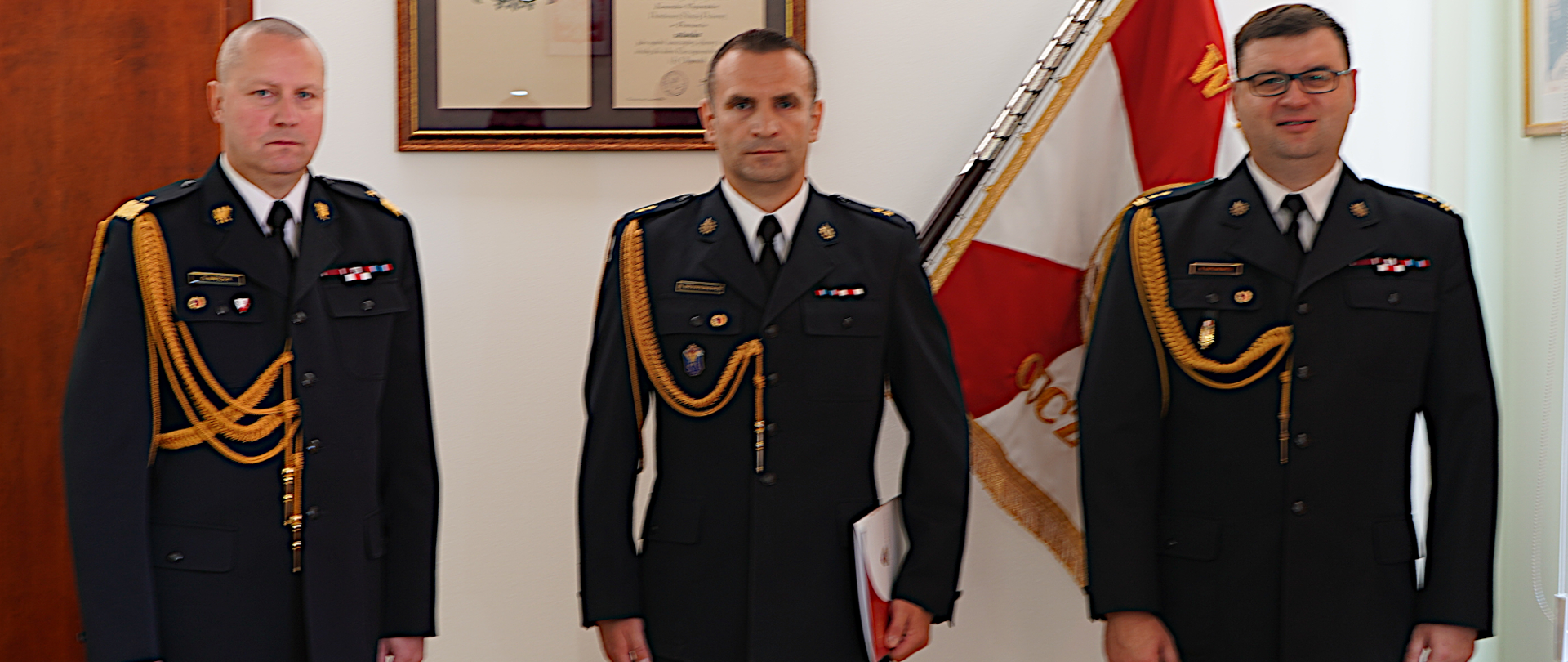 Trzech funkcjonariuszy stoi ubranych w mundury koloru grafitowego jeden w stopniu generalskim z niebieskim i lampasami za nim na ścianie sztandar koloru biało-czerwonego