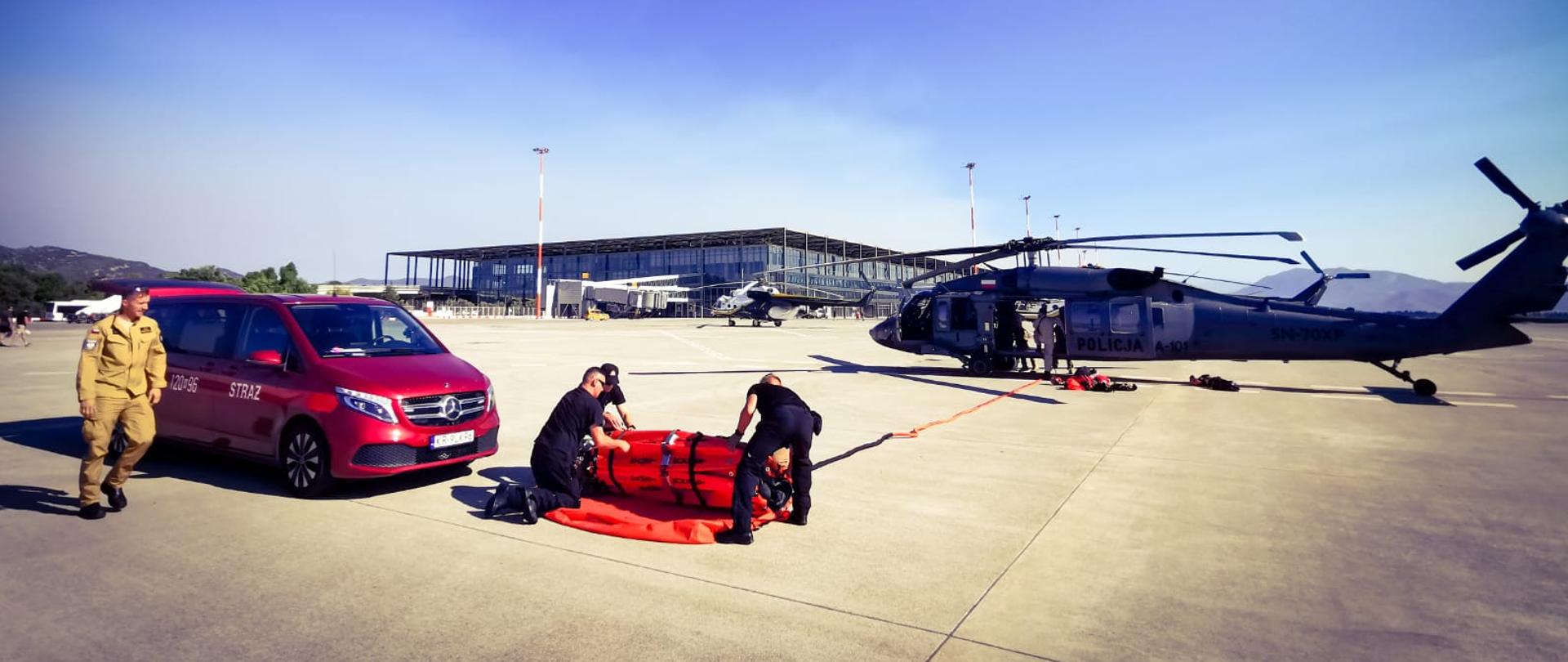 Strażacy instalujący na płycie lotniska zbiornik typu "Bambi Bucket. do śmigłowca. Z prawej strony widoczny pojazd straży pożarnej
