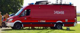 Czerwony samochód dostawczy straży pożarnej (Mercedes-Benz Sprinter) z białą zderzakową częścią przednią i numerem taktycznym „340 P 98” na bocznej ścianie. Na dachu pojazdu zamontowane są dwa wysokie, pionowe maszty antenowe z owiniętymi okablowaniami. Pojazd stoi na terenie zielonym, po lewej stronie w tle widoczni są strażacy w piaskowych mundurach oraz fragmenty innych samochodów.