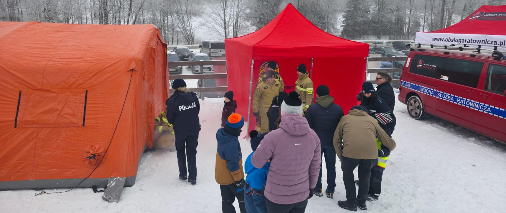 Grupa uczestników pokazów strażackich na tle rozstawionych namiotów. Funkcjonariusze omawiają zasady bezpiecznego wypoczynku.