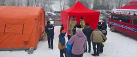 Grupa uczestników pokazów strażackich na tle rozstawionych namiotów. Funkcjonariusze omawiają zasady bezpiecznego wypoczynku.