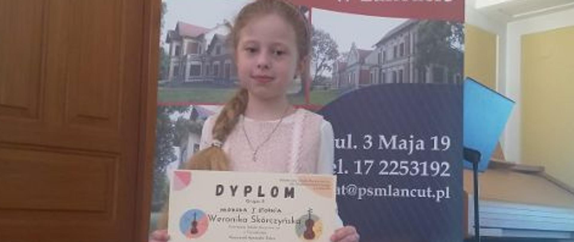 Zdjęcie przedstawiające uczennicę w różowej sukience trzymającą dyplom.