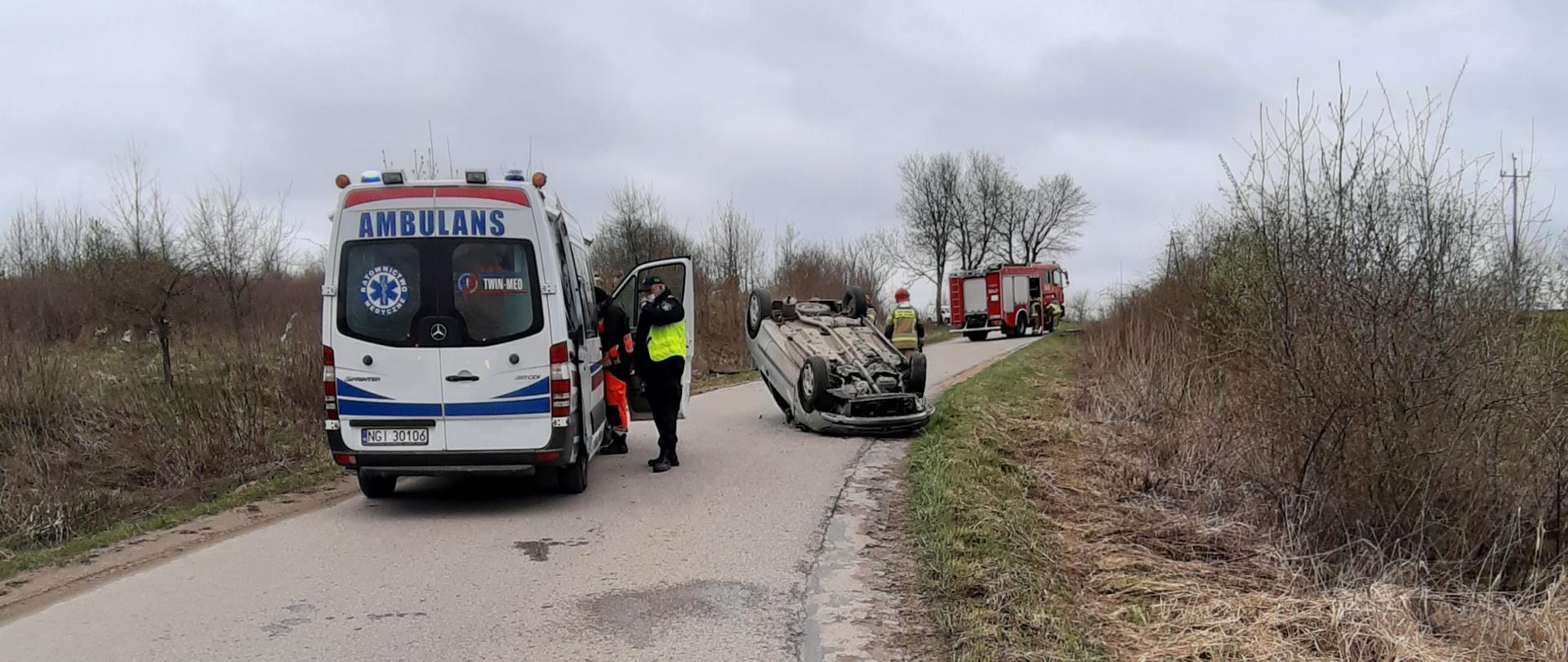 Na drodze znajduje się samochód osoby, który leży na dachu. Przed pojazdem stoi ZRM, przy którym stoi policjant i ratownik medyczny. Z kolei za pojazdem leżącym na dachu stoi samochód straży pożarnej 